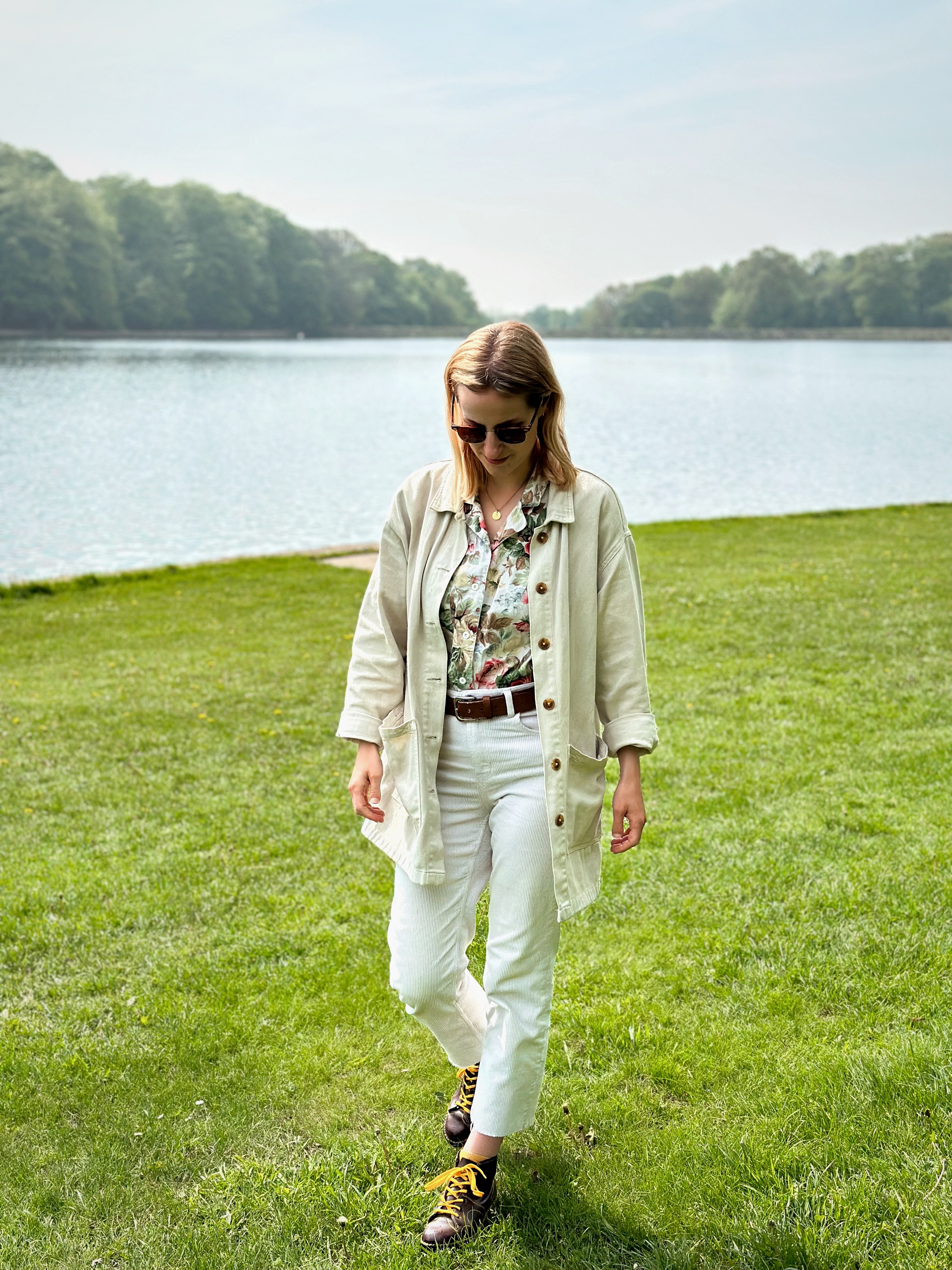 Amy is walking in front of a lake, looking down. She is wearing cream trousers and a cream oversized jacket.