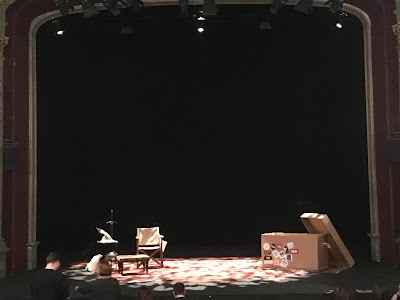 Stage in darkness, in foreground , a table with Gandalf's wizards hat, a directors chair, and a large cardboard crate decorated with stickers 