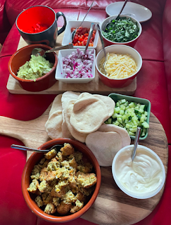 A photo of a tray of small bowls. Each one is filled with taco ingredients, such as chopped peppers, coriander, red onion, cucumber, tomatos, avocado, grated cheese, sour cream, and crumbled falafel. There are mini tacos on the side ready to be made.