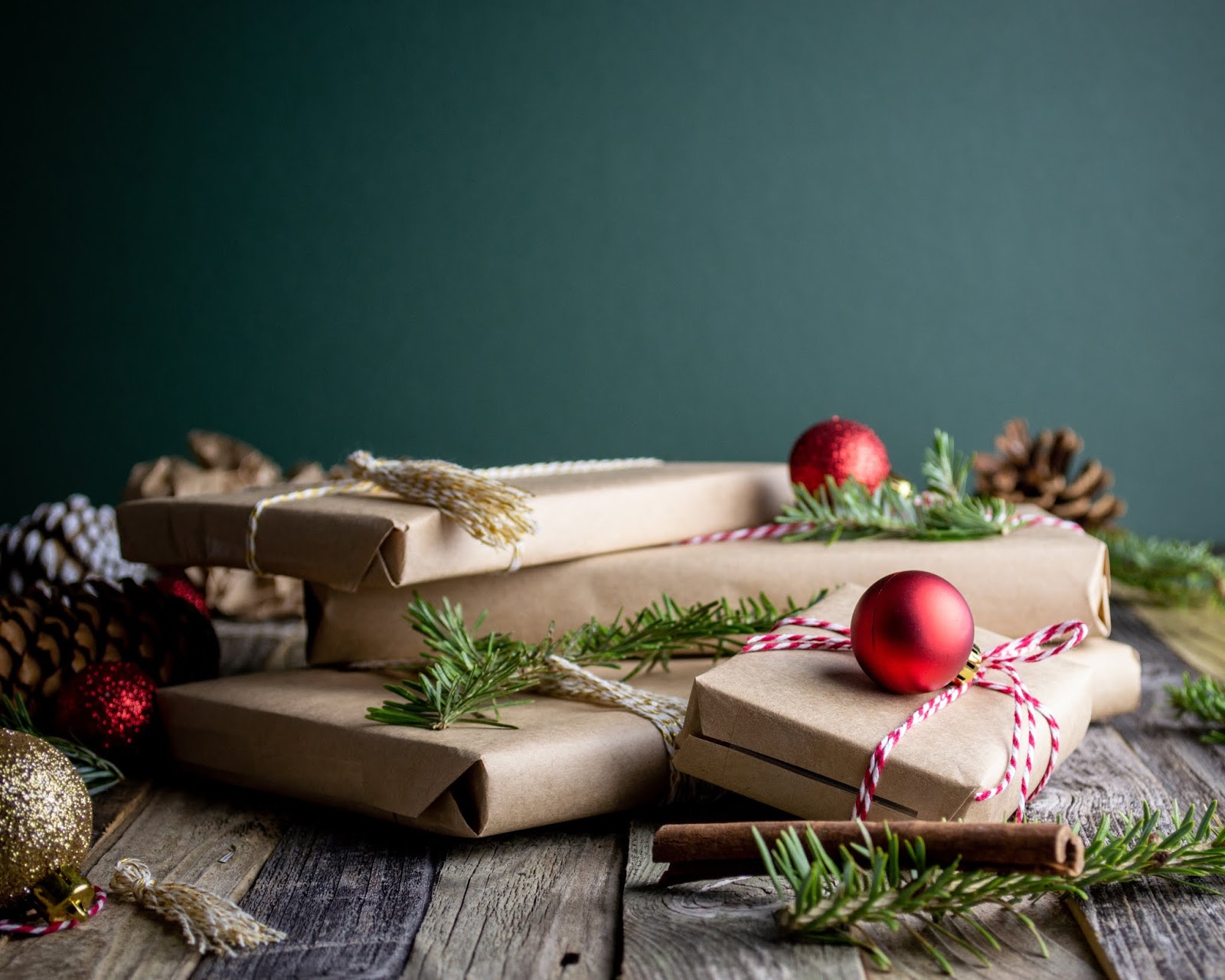 Four presents wrapped in brown paper with red and white ribbon string around them, in front of a teal background, surrounded by sprigs of pine, baubles, and cinnamon sticks.