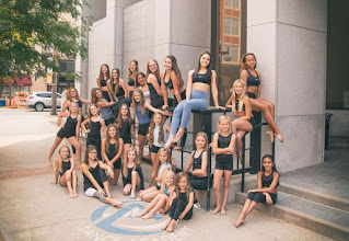 young dancers in various black dance costumes lean against a downtown building and smile