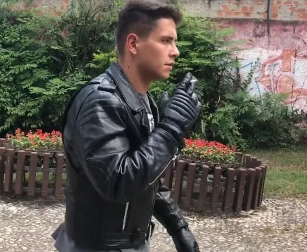 Side view from the waist up a man smoking a cigarette wearing a black leather biker jacket outside in a park