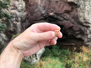 A photo of a small earthenware skull (Skulferatu 84) being held up in front of the entrance to St Baldred's Cave.  Photograph by Kevin Nosferatu for the Skulferatu Project.