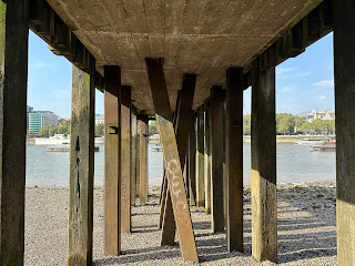 A view under the wooden structure of Gabriel's Pier looking down the beach towards the waters of the Thames.  There are wooden posts on both sides with the floor of the pier above.  In the middle there are several posts standing at angles against each other.  Photo taken by Kevin Nosferatu for the Skulferatu Project.