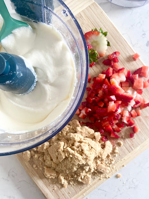 Food processor with creamy ice cream + diced strawberries and crushed graham crackers on a wooden cutting board.