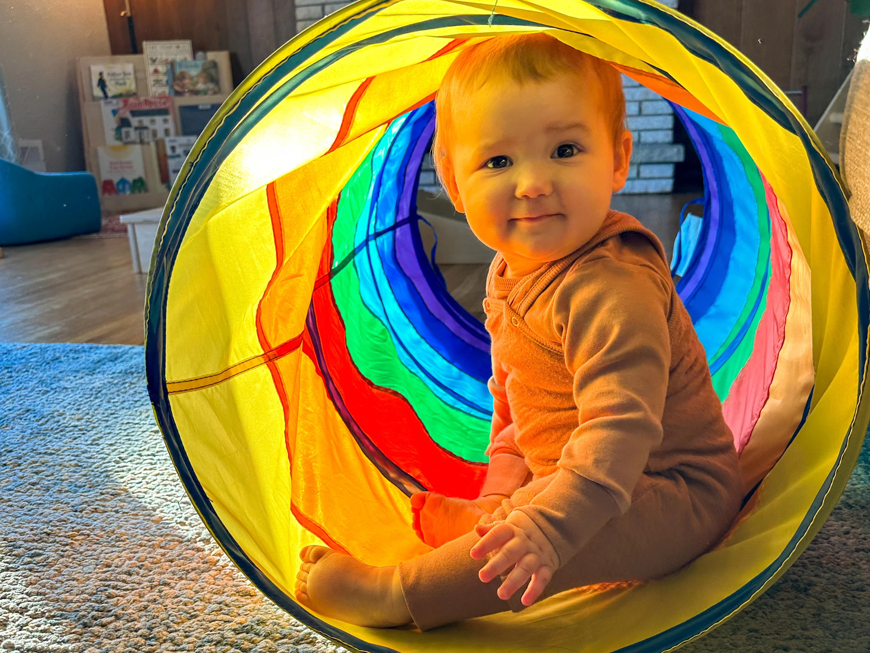 Baby sits in play tunnel in Montessori home while smiling.