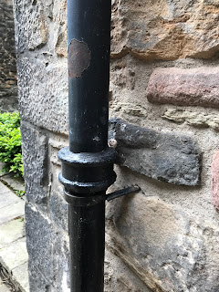 A photo of a small, ceramic skull (Skulferatu #83) resting in the curve of an old black drainpipe at the side of the doorway into Acheson House.  Photo by Kevin Nosferatu for the Skulferatu Project.