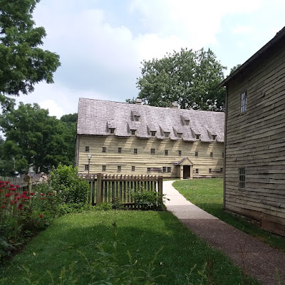 Two-story wooden sided medieval style building with roof dormers. A curved path runs from the building to the front of the photo through a green lawn and there is a wooden fence with flowers planted along it.