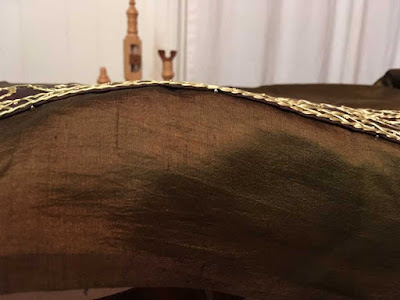 Green-gold silk lifted to show the edge of a gold-embroidered strip of brown fabric, with occasional silver pin heads showing just under the folded trim edge.