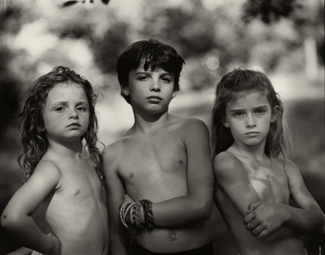 A foto em preto e branco retrata três crianças do quadril para cima, as três sem camisa e de fisionomia séria. A menina à esquerda, mais nova que os outros dois, está com as mãos na lateral do corpo e cabelo solto. No centro temos um menino que parece ser o mais velho, e à direita há outra menina, com os braços cruzados e a frente do cabelo preso.