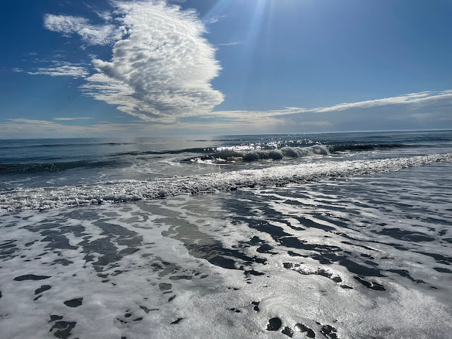 The first of three pictures that shows the progression of a wave as it comes into the shore, crests, and hits the shoreline. There is a single cloud that is all by itself above where this wave is building. It is a weird shaped cloud. It looks like smoke pouring out of a chimney stacked fluffy and white. The wave I was following is just building in this photo.