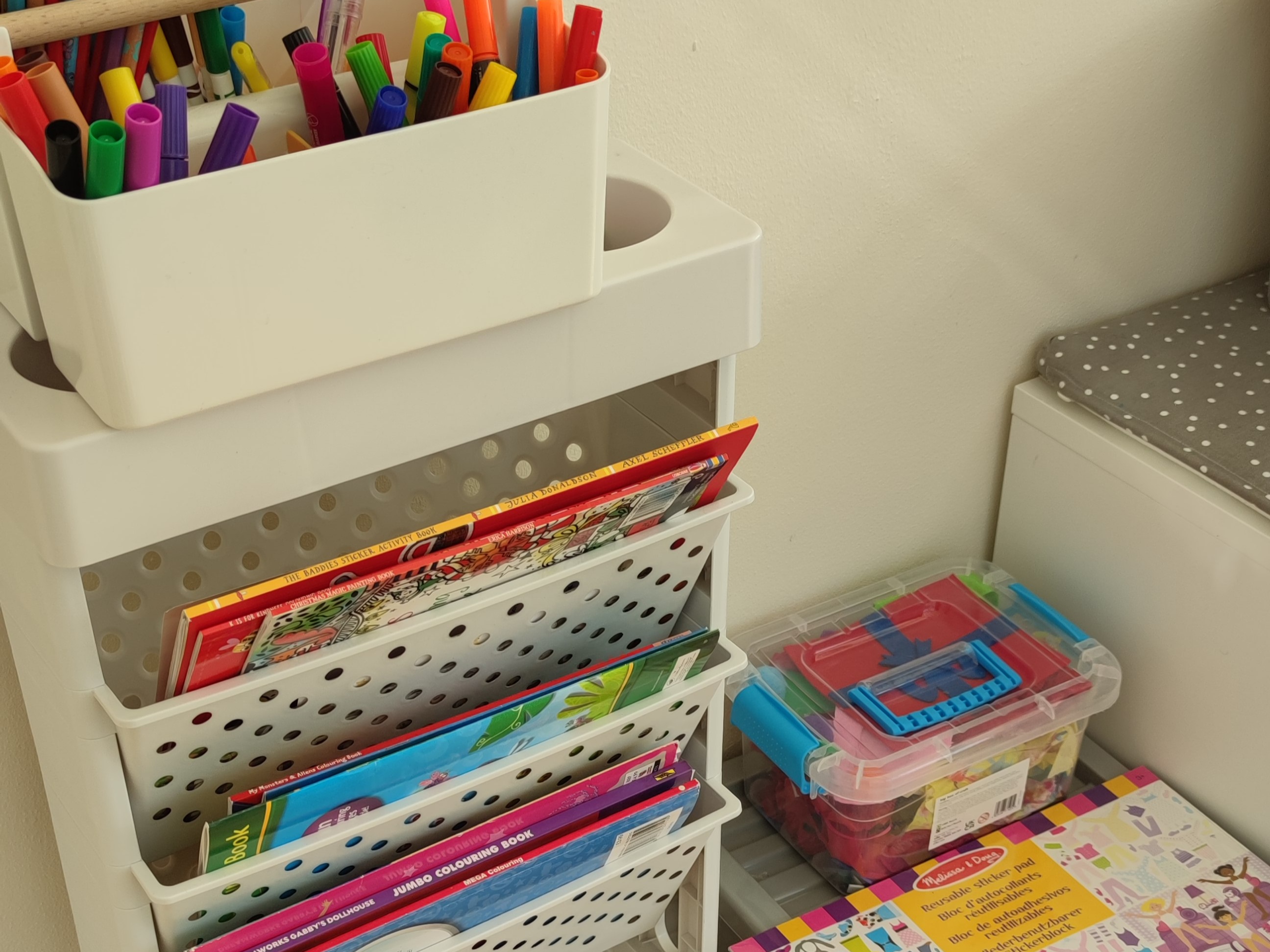 Storage shelves with colourful art supplies and a storage caddy containing colouring books and educational materials, arranged next to a dotted cushion.
