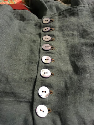A column of eight blue-iridescent two-hole shell buttons, stitched on with tan thread and done up through tan buttonholes, in a muted-teal skirt yoke.