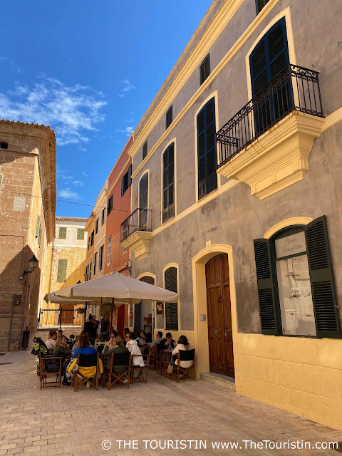 People sitting on wooden and dark grey director chairs at wooden bistro tables under large white sun umbrellas on a square lined by pastel-coloured period houses in front of a restaurant.