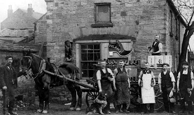 Seven male workers stood outside the Wm Lamb Clog Manufactuers Shop. There is a horse and cart with an array of clogs on display, and one of the men is stood on the cart holding a pair for sale. The other workers hold an array of tools and clogs, and one has his hand petting a dog.