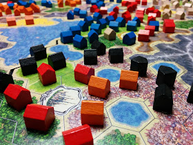 A close-up view of a game after it has ended. There are four sections of the game board, each placed together to form the play area. Each one is made up of a grid of hexagonal spaces, coloured to represent grasslands, forests, flowers, canyons, mountains, lakes, and deserts, with a fancy space representing a city. On most of the non-water spaces is a wooden house token in one of five colours: blue, white, black, red, and orange.