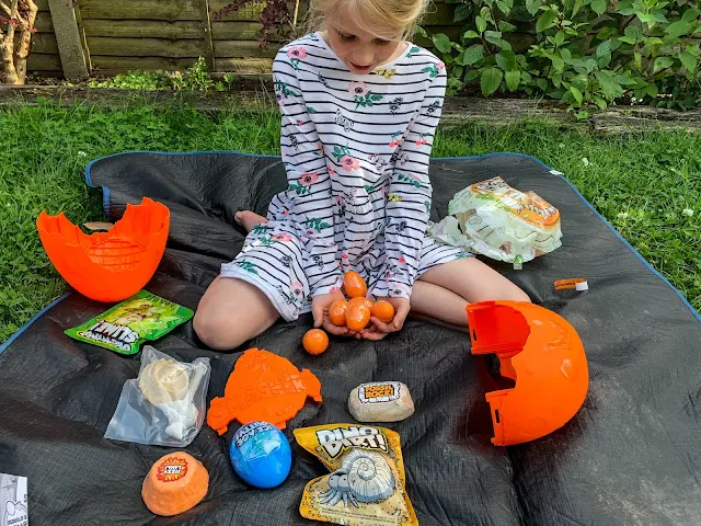 A girl showing 6 orange Smashers eggs and surrounded by the contents of the Epic Dino Egg including the egg shell, ice age putty, fizzy lava, glowing slime, dino dirt and fossil rock