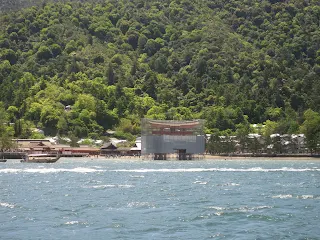 View of giant torii gate on Miyajima island from the water. The ocean is in the foreground and the background is a large wooded mountain. A very large red torii gate can be seen in the water covered in nets and scaffolding