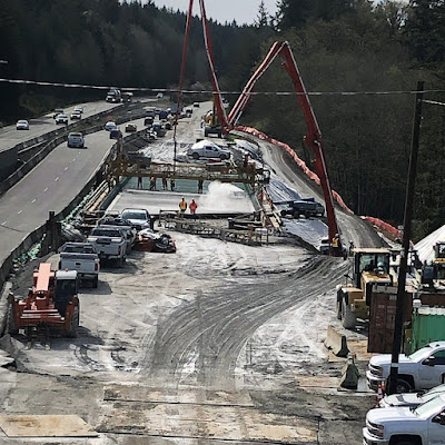 Photo of work zone with large cranes and work trucks around a new bridge under construction where workers pour concrete onto the bridge deck.