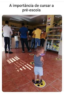 Foto. Em uma padaria/cafeteria, adultos e crianças em pé, posicionados em uma fila mal organizada. No chão de lajotas quadradas avermelhadas, a sinalização com distanciamento ignorado pelas pessoas. Distante do balcão e sozinho, um garotinho, pisa sobre uma das marcas redondas amarelas estampadas em preto em forma de solado. Ele está de costas, bracinhos cruzados e olha à direita; tem cabelos castanhos curtos  bem  finos, usa máscara colorida, regata cinza escuro, bermuda de malha cinza claro e sandálias pretas.  No topo lê-se: A importância de cursar a pré-escola.