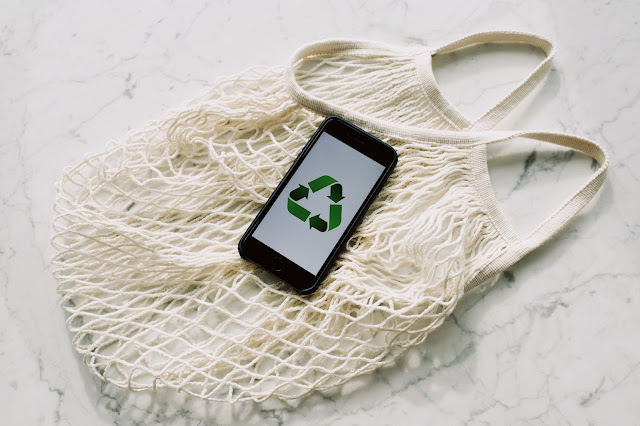 There is a white marble table with a white netted shopping back on top. The bag is reusable. In the centre of the image, on top of the bag, there is a black iphone which has the recycling image on the screen