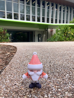 Tiny Santa stands outside the Southern Entrance of the Eddie Koiki Mabo Library. He is a plastic Santa figurine with articulated joints and an oversized head, that was made on a 3D printer using white plastic filament. He has been hand coloured with sharpies, but his clothing is a light reddish-orange colour rather than a bright dark red and the overall affect is charming, rather than tacky.