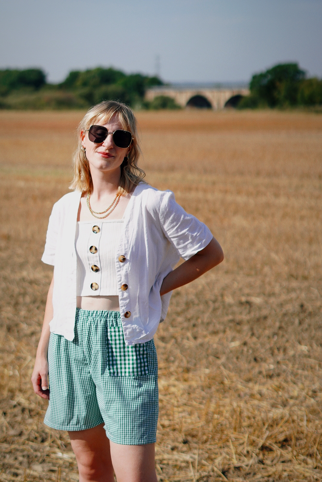 Amy is stood in a wheat field on a sunny day, hand on one hip looking to camera. She is wearing a white shorty sleeved shirt, green gingham shorts and sunglasses.