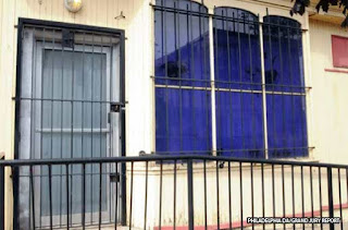 Color photograph showing a railed entrance to a door blocked by a heavy security gate with a large lock. Three security barred windows are to the right of the door.