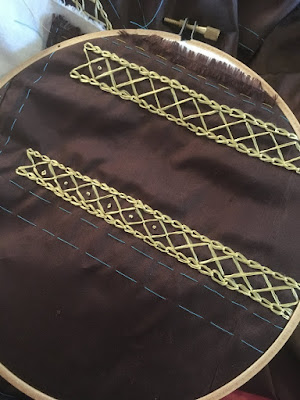 Dark brown silk stretched in a wooden embroidery hoop, with two narrow bands of diamond stitches bordered with chain stitch lines, and French knots starting back down the rows of diamonds.