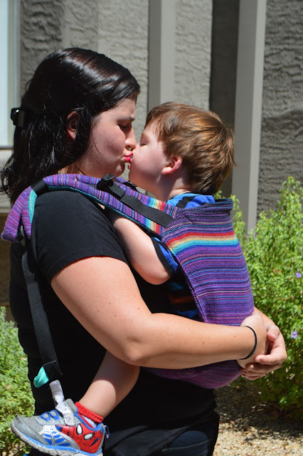 Image of fair skin woman with dark hair kissing fair skin brown haired toddler worn in a front carry in a purple onbu with rainbow stripes