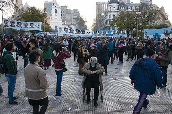 persona mayor en silla de ruedas, manifestando en la plaza de mayo con la bandera de los próceres argentinos de fondo