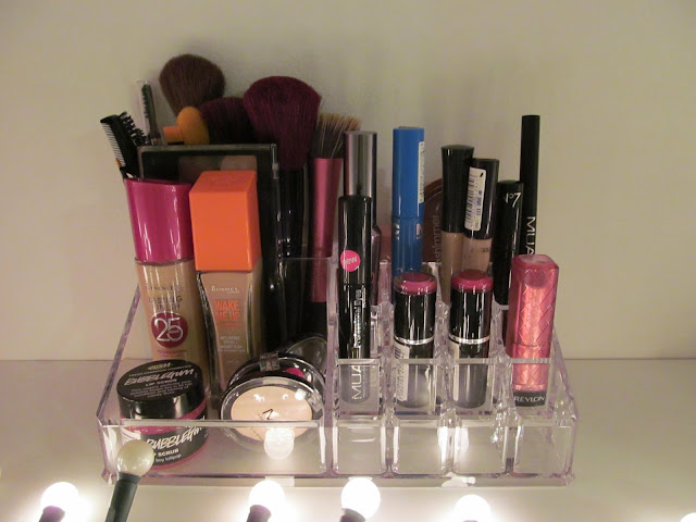 acrylic makeup storage lit by fairy lights in the foreground containing several lipsticks, two bottles of foundation and various tubes and pots