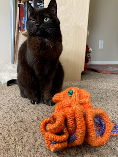 Tinycat (small, black, permanent resting bitch face) pretends to ignore the vivid orange knit octopus sitting at her feet.