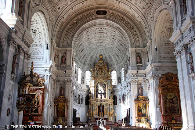 A large golden-coloured altar and a large oil painting in light green and blue colours in an elaborately decorated overall white interior of a church with white stone columns, and statues, angels, and dark brown wooden benches, under a vaulted elaborately decorated white ceiling.