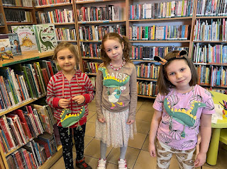 Trzy dziewczynki stoją na tle regałów z książkami  w bibliotece. Dziewczynka po lewej trzyma w ręce pracę plastyczną: papierowego dinozaura. Dziewczynka stojąca w środku i po prawej stronie prace plastyczną powieszoną mają na szyi.