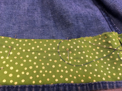 A close up of a narrow strip of dark lime-green fabric with scattered gold dots, stitched into the hem of a denim skirt and pinned at the top, folded under edge.