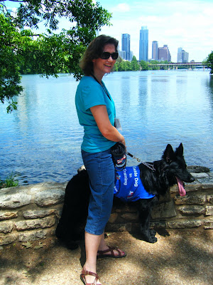 Petey, big black German Shepherd, and I are standing in front of a low stone wall; he's in his work jacket; the water of Town Lake is behind us and the Austin skyline in the background