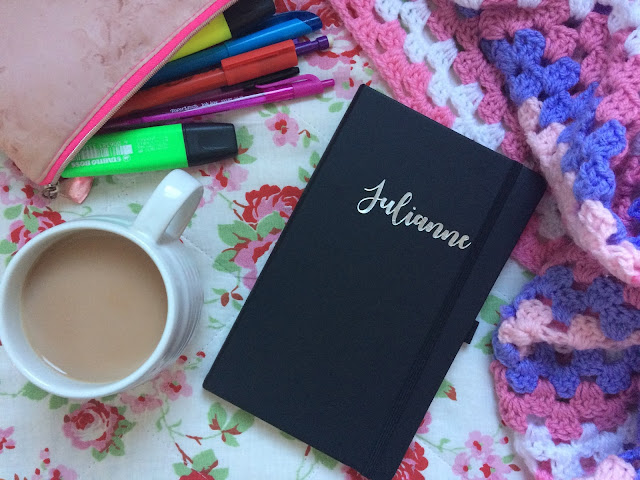 Black notebook with silver 'Julianne' title, cup of tea in white mug, pink pencil case with colourful pens and highlighters spilling out, pink purple and white crochet blanket on right