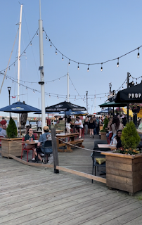 The waterfront at Halifax. There are a selection of outside restaurants and cafes. The sky is blue, and the sea is in the distance. It's clearly a warm day as people are wearing summer clothes. It feels relaxing just to look at it. There are sun umbrellas up at tables and a string of fairy lights between posts.