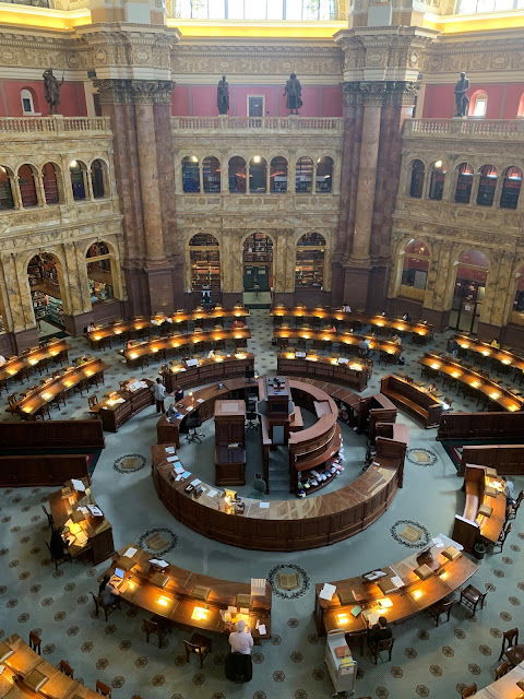 A circular desk with shelves in the middle sits in the middle of the room. Long straight desks with lights and people sitting at them circle this. Statues and archways surround the outside of the room.