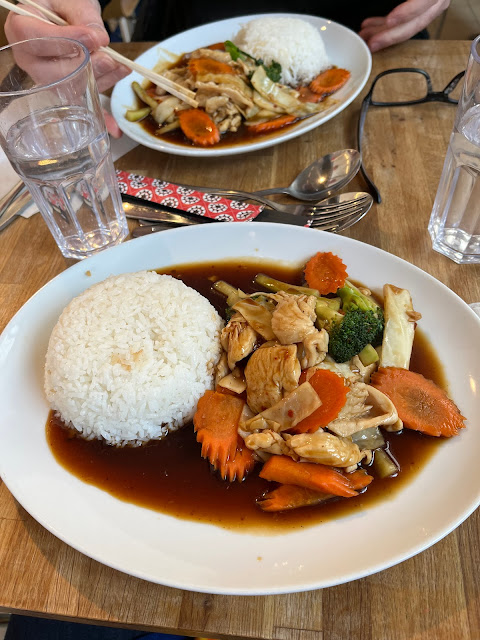 A white oval plate with a chicken, carrot and broccoli dish, a mound of white rice, glass of water and chopsticks on a wooden table