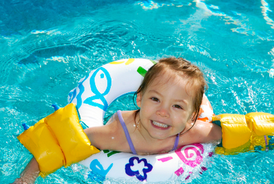 Picture of a happy  little girl in the pool wearing floaties and float ring. Confidence, when you learn how to swim, is always very important in new swimmers.