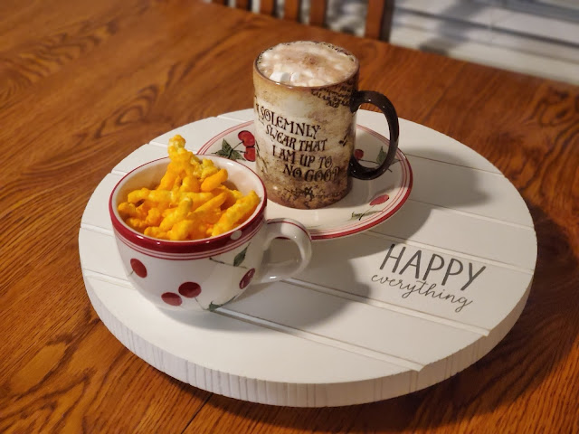 White Lazy Susan that says "happy everything" in black lettering, on top of a wooden table. On top of the Lazy Susan is a soup bowl with cheetos inside and a Harry Potter coffee cup that says "I solemnly swear I am up to no good." The coffee cup is full to the brim with hot chocolate and mini marshmallows