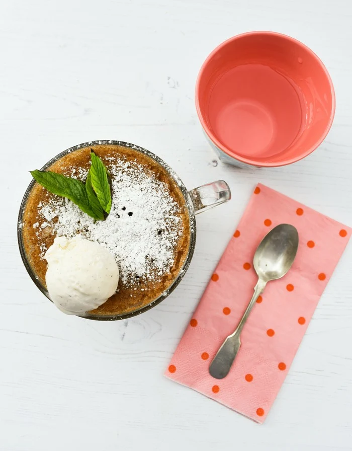 Overhead shot of gingerbread cake in a mug next to pink spotty napkin and a teaspoon