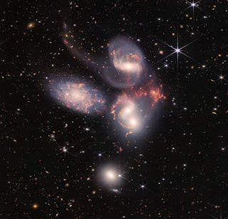 Stephan’s Quintet (NIRCam + MIRI Imaging) - NGC 7317, NGC 7318A, NGC 7318B, NGC 7319, and NGC 7320. A group of five galaxies that appear close to each other in the sky: two in the middle, one toward the top, one to the upper left, and one toward the bottom. Four of the five appear to be touching. One is somewhat separated. In the image, the galaxies are large relative to the hundreds of much smaller (more distant) galaxies in the background. All five galaxies have bright white cores. Each has a slightly different size, shape, structure, and coloring. Scattered across the image, in front of the galaxies are number of foreground stars with diffraction spikes: bright white points, each with eight bright lines radiating out from the center.