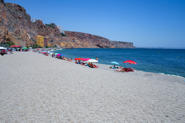 Amplia playa con forma de bahía y acantilados con sombrillas de colores y turistas al fondo