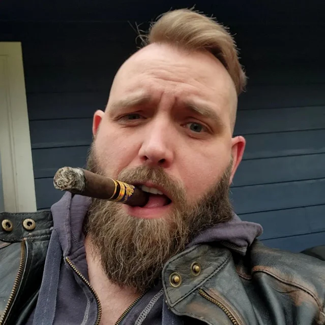 11/12 some shoulders up a handsome blonde bearded bear wearing brown leather jacket smoking a stogie