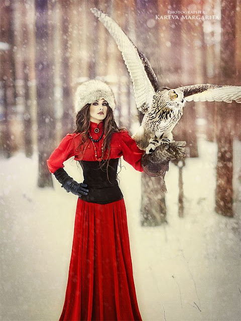 Woman wearing Neo-Victorian clothing (fur hat, underbust corset, red blouse, red skirt, gloves) in the snow with an owl perched on her glove