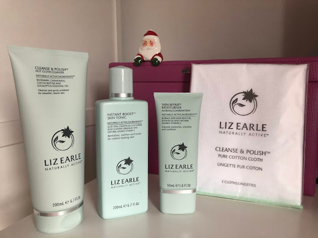 Liz Earle cleanser, toner and moisturiser pale green bottles next to white cotton cloths with small Santa ornament behind