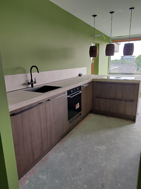 Keuken met eikenhouten kleurige keukenkasten, zwarte kraan, zwarte spoelbak en een licht beige aanrechtblad. Drie tonnetjes aan een ketting als lamp
